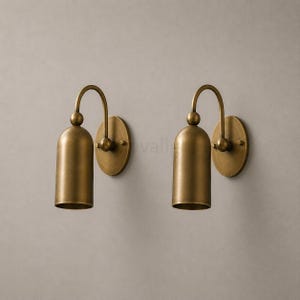 Könnte beinhalten: Ein Paar Wandleuchten mit gebürstetem Messing-Finish. Jede Leuchte besteht aus einer runden Wandplatte, einem geschwungenen Arm mit kugelförmigem Akzent und einem zylindrischen, nach unten gerichteten Lampenschirm mit offener Unterseite.