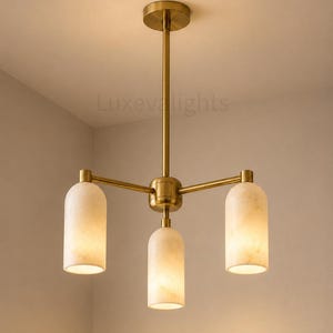May include: A three-light chandelier featuring a brushed brass finish central rod and frame with three cylindrical, off-white marble shades. The fixture includes a circular ceiling canopy and the text Luxevelights is visible in the background.