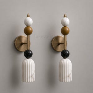 May include: Two wall sconces with a modern design. Each sconce features a brass base, a vertical rod with a white marble sphere, a brown sphere, a black sphere, and a white marble, fluted base. The sconces are mounted on a gray wall.