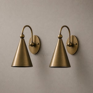 May include: A pair of wall sconces featuring a brushed brass finish. Each light fixture consists of a circular wall-mounted plate, a curved arm, and a downward-facing conical shade. The metal surface has a matte, metallic texture throughout.