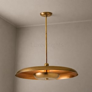 May include: A brass pendant light fixture featuring a long, thin vertical rod connected to a wide, flat, circular shade. A smaller, bowl-shaped brass component is centered beneath the main shade, creating a layered, metallic design.