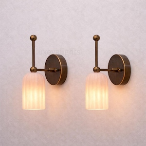 Può includere: Due applique da parete con una luce calda e accogliente. Ogni applique presenta un paralume cilindrico, scanalato, bianco sporco, una piastra posteriore circolare color bronzo e un braccio verticale sottile. Il design è semplice ma elegante.