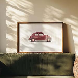May include: A framed print featuring a maroon vintage car illustration. The car has a rounded body, small wheels, and a classic design. The artwork is set against a white background and framed in a wooden frame. The print is displayed on a green velvet sofa.