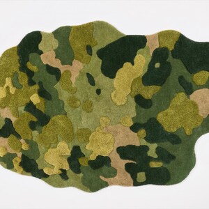 May include: A camouflage-patterned rug in shades of green, tan, and yellow. The rug has an irregular, organic shape and a textured surface. The design features a mix of solid and mottled areas, creating a visual effect.