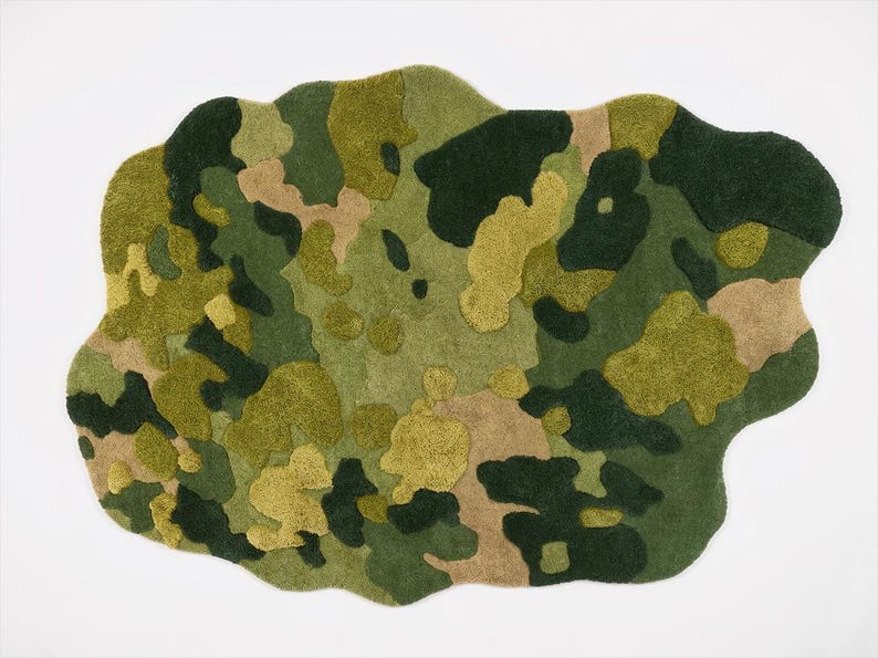 May include: A camouflage-patterned rug in shades of green, tan, and yellow. The rug has an irregular, organic shape and a textured surface. The design features a mix of solid and mottled areas, creating a visual effect.