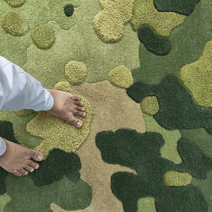May include: A green and tan camouflage-patterned rug with a textured, tufted design. The rug features various shades of green and tan, creating a camouflage effect. The design includes raised, rounded shapes. The rug is made of a soft material.