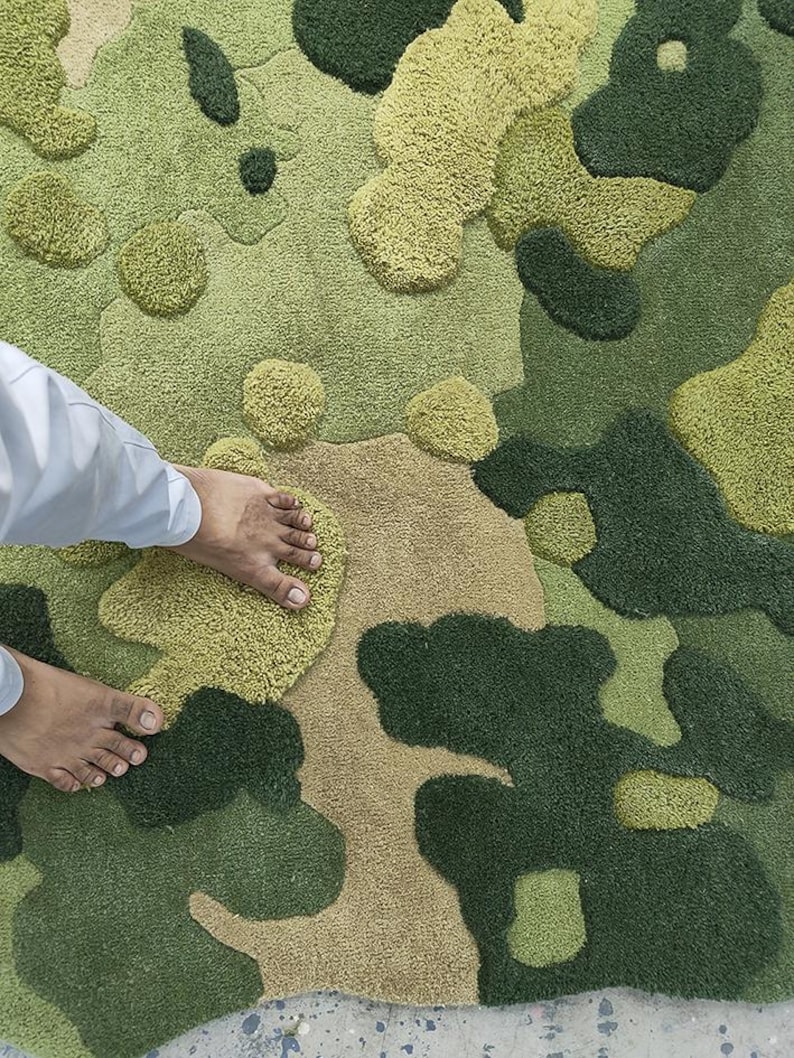May include: A green and tan camouflage-patterned rug with a textured, tufted design. The rug features various shades of green and tan, creating a camouflage effect. The design includes raised, rounded shapes. The rug is made of a soft material.