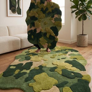 May include: A camouflage-patterned rug in shades of green, tan, and olive. The rug has an irregular, organic shape and a textured, tufted surface. The rug is displayed on a hardwood floor.