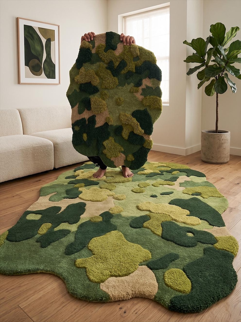 May include: A camouflage-patterned rug in shades of green, tan, and olive. The rug has an irregular, organic shape and a textured, tufted surface. The rug is displayed on a hardwood floor.