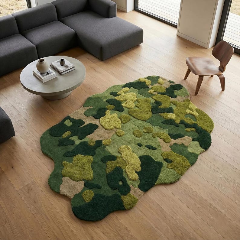 May include: A green and beige camouflage-patterned rug with an irregular shape. The rug features varying shades of green, creating a textured, three-dimensional effect. The rug is placed on a light-colored wooden floor, with a gray sectional sofa and a round coffee table in the background.