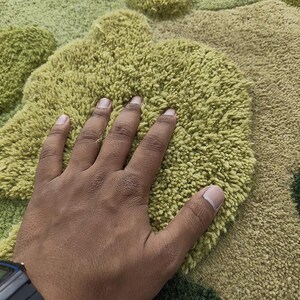 May include: A close-up of a textured rug with a hand resting on it. The rug features a pattern of green and tan shapes. The rug's texture appears soft and plush.