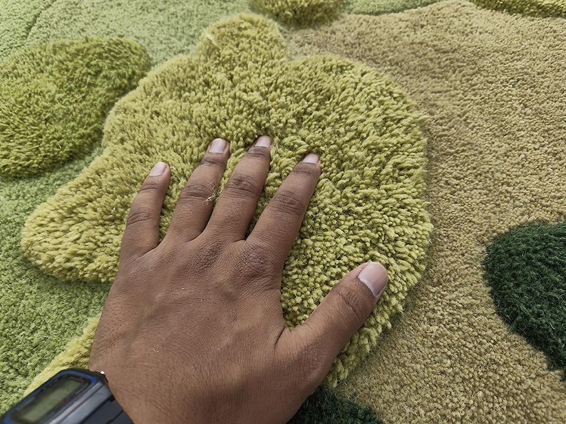 May include: A close-up of a textured rug with a hand resting on it. The rug features a pattern of green and tan shapes. The rug's texture appears soft and plush.