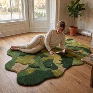 May include: A unique, abstract rug in shades of green, tan, and olive. The rug has an irregular, organic shape and a textured surface. The rug is placed on a hardwood floor in a well-lit room.