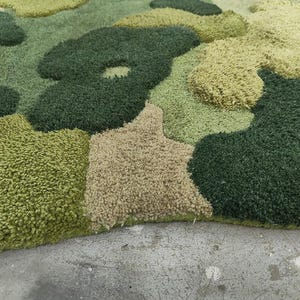 May include: A close-up of a green, beige, and olive-colored rug with a camouflage pattern. The rug has a textured, plush surface and is made of a soft material. The rug's design features irregular shapes and varying shades of green and beige.