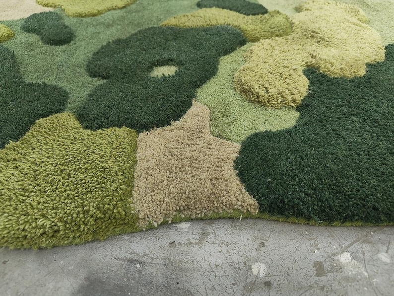 May include: A close-up of a green, beige, and olive-colored rug with a camouflage pattern. The rug has a textured, plush surface and is made of a soft material. The rug's design features irregular shapes and varying shades of green and beige.