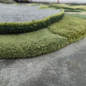 May include: Close-up of a green and gray rug with a textured, plush surface. The rug features a layered design with varying shades of green and a gray backing. The edges are neatly finished with a dark green border.