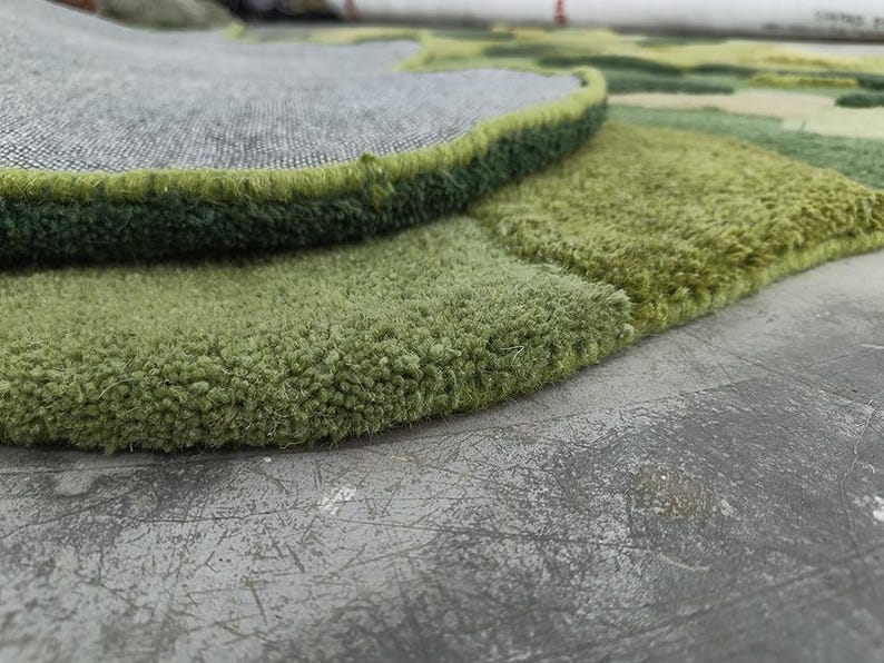 May include: Close-up of a green and gray rug with a textured, plush surface. The rug features a layered design with varying shades of green and a gray backing. The edges are neatly finished with a dark green border.