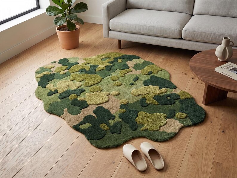 May include: A camouflage-patterned rug in shades of green, beige, and brown, with an irregular, organic shape. The rug is placed on a hardwood floor, with a sofa, coffee table, and plant visible in the background. A pair of cream-colored slippers are on the floor.