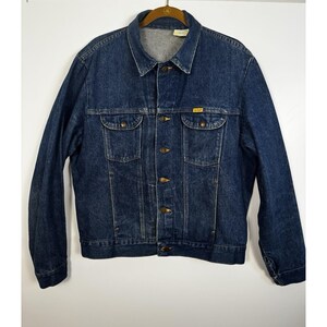 May include: A dark blue denim jacket featuring a pointed collar, long sleeves, and a button-up front. The jacket includes two chest pockets with buttoned flaps and a small yellow rectangular brand tag on the left chest pocket seam.