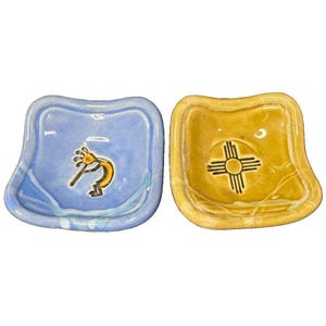 May include: Two small, square ceramic dishes with rounded corners. One dish is light blue with a dark yellow Kokopelli figure in the center. The other dish is mustard yellow with a dark brown Zia sun symbol centered on the glazed surface.