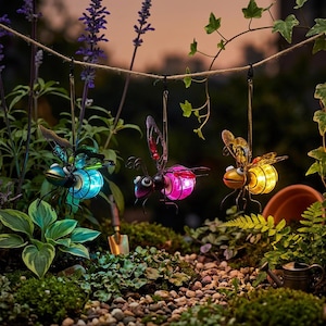 May include: Three decorative metal insect garden lights hanging from a string. The insects have colorful, glowing bodies in blue, pink, and yellow. The garden setting includes plants, rocks, and a watering can, creating a whimsical outdoor scene.