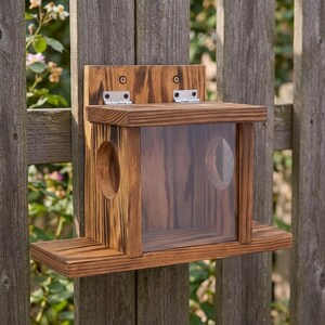 Könnte beinhalten: Ein hölzernes Vogelhaus mit einer transparenten Frontplatte und zwei kreisförmigen Eingangsöffnungen. Das Vogelhaus ist an einem Holzzaun befestigt. Es hat eine kleine Sitzstange und einen aufklappbaren Deckel.