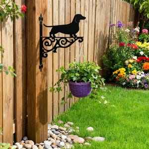 May include: Black metal dachshund dog silhouette plant hanger with decorative scrollwork. A purple woven basket with green plants hangs below. The hanger is mounted on a wooden fence post in a garden setting.