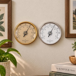 Op de afbeelding: Twee ronde analoge thermometers naast elkaar. De ene heeft een goudkleurige rand met een beige wijzerplaat, de andere een zilverkleurige rand met een witte wijzerplaat. Beide tonen dubbele temperatuurschalen in Fahrenheit en Celsius.
