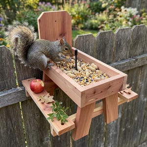 Può includere: Un alimentatore per scoiattoli in legno a forma di tavolo da picnic, pieno di arachidi e semi. Uno scoiattolo grigio mangia dall'alimentatore. Una mela e delle foglie sono sulla panca attaccata. L'alimentatore è montato su una staccionata di legno.