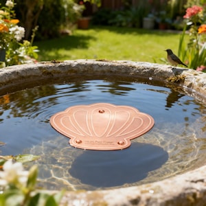 May include: A copper bird bath water protector shaped like a stylized shell, floating in a stone bird bath filled with water. The protector has the text "USA Pure Copper" and is surrounded by a lush green garden.