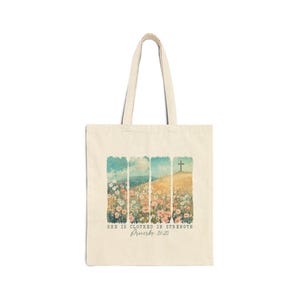 May include: A cream-coloured canvas tote bag featuring a vertical panel graphic of a wildflower meadow and a hill with a cross. Below the illustration, the text reads: SHE IS CLOTHED IN STRENGTH Proverbs 31:25 in a dark, serif font.