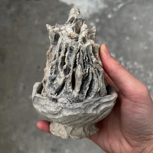 Puede incluir: Un quemador de incienso de cerámica esculpido a mano con un acabado gris rugoso y texturizado. El objeto presenta una base ancha en forma de cuenco y una parte superior hueca en forma de cono con aberturas verticales que revelan un interior oscuro.