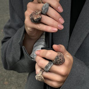 May include: A collection of six rings with irregular, textured surfaces in shades of gray, brown, and tan. Each ring features a unique, organic shape with deep crevices and raised details, worn across multiple fingers on both hands.