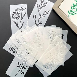 Op de afbeelding: Een verzameling bloemen sjablonen in verschillende ontwerpen, waaronder zonnebloemen, bladeren en takken. De sjablonen zijn gemaakt van een doorschijnend materiaal en zijn gerangschikt op een donkere ondergrond. Een ingelijst kunstwerk met een groen bloemenontwerp is zichtbaar op de achtergrond.