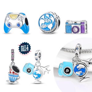 May include: Collection of silver charms with travel and gaming themes. Charms include a blue and silver video game controller, a globe with "Let's Go" text, a blue and purple camera, and travel-themed charms.