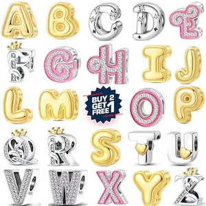 Puede incluir: Una colección de dijes con letras del alfabeto para pulseras, con acabados en tono dorado, plateado y esmalte rosa con detalles de cristal. Algunas letras incluyen motivos de corona o corazones. Un gráfico central azul dice BUY 2 GET 1.