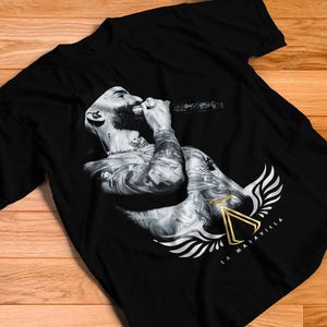 Op de afbeelding: Een zwart t-shirt met korte mouwen en een monochrome afbeelding van een persoon met een baard die in een microfoon zingt. Het ontwerp bevat een goudkleurig driehoekig embleem met vleugels en de tekst LA MARAVILLA onder de afbeelding.