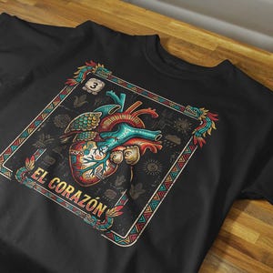 May include: A black t-shirt featuring a graphic print of an anatomical heart with colorful patterns. The design is framed by a decorative border with serpent motifs and the text EL CORAZÓN in yellow letters, along with the number 3 in a small box.