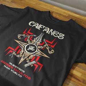 Könnte beinhalten: Ein schwarzes kurzärmeliges T-Shirt mit einem Grafikdruck, der oben den Text CAIFANES und unten NUESTROS DIOSES OCULTOS HAN VUELTO zeigt. Das zentrale Design umfasst einen Stern, Gitarren und rote geometrische Muster auf schwarzem Stoff.