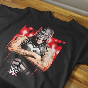 May include: A black short-sleeved t-shirt featuring a graphic print of a masked wrestler with extensive arm tattoos. The wrestler is wearing a black mask with white star details and has their tongue out. The WWE logo is printed in white at the bottom.