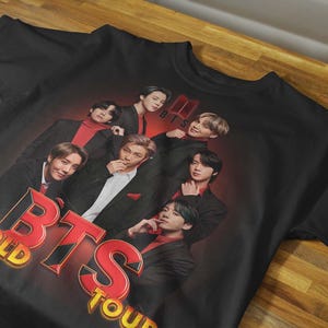 May include: A black short-sleeved t-shirt featuring a graphic print of the seven members of the music group BTS. The design includes the red and yellow text "BTS WORLD TOUR" and the BTS logo, set against a dark red and black gradient background.