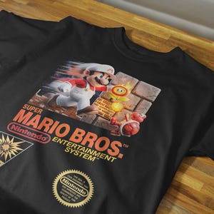 May include: A black short-sleeve t-shirt featuring a graphic of Mario from Super Mario Bros. running past a fire flower and a red turtle shell. The text reads Super Mario Bros. Entertainment System, Nintendo, Action Series, and a gold seal.