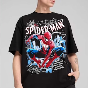Peut inclure: Un t-shirt noir à manches courtes avec un motif représentant Spider-Man accroupi devant une ligne d'horizon urbaine et une toile d'araignée. Le texte indique "THE SPIDER-MAN" en haut et "With great power comes great responsibility" en bas.
