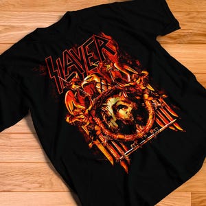 May include: A black short-sleeved t-shirt featuring a graphic print of the band Slayer logo in red text above an illustration of a circular emblem with a skull, surrounded by sharp sword-like shapes and fiery orange and yellow flame accents.