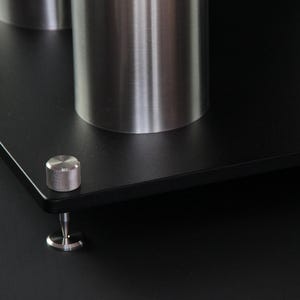 Könnte beinhalten: Nahaufnahme einer schwarzen, rechteckigen Plattform mit drei zylindrischen, gebürsteten Metallstützen. Ein kleiner, silberner Knopf und ein Fuß sind an der Ecke sichtbar. Die Plattform ist für die Aufnahme von Objekten konzipiert.