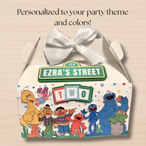 May include: A white cardboard party favor box topped with a white satin ribbon bow. The box features a green street sign reading "EZRA'S STREET" and a sign reading "TWO" above colorful Sesame Street character cutouts on a polka dot background.