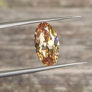 Pode incluir: Uma gema ovalada castanho-dourada é segurada por pinças prateadas contra um fundo de madeira desfocado. As facetas da gema refletem a luz, criando um efeito cintilante. As pinças estão em foco.