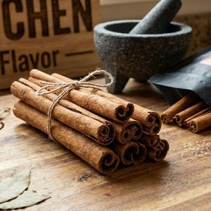 May include: A bundle of brown cinnamon sticks tied with twine, positioned next to a black pouch labeled Chuco Kitchen Whole Cinnamon Sticks. Nearby are dried red chili peppers, bay leaves, a stone mortar and pestle, and a small bowl of brown sugar.