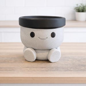 May include: A gray ceramic planter shaped like a smiling character with a black rim and black eyes. The planter has a round body with small feet and is sitting on a wooden surface. The background is a white brick wall.