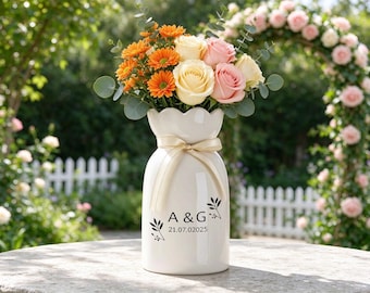 May include: A white ceramic vase with a scalloped rim and a cream-coloured ribbon tied around the neck. The vase features black text reading A & G and the date 21.07.2025, accented by small leaf illustrations, filled with orange daisies and pink roses.